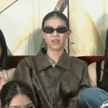 a woman wearing sunglasses and a brown jacket is sitting on a couch with other women .