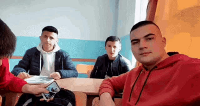 a group of young men are sitting at a table in a classroom and one of them is holding a cell phone .