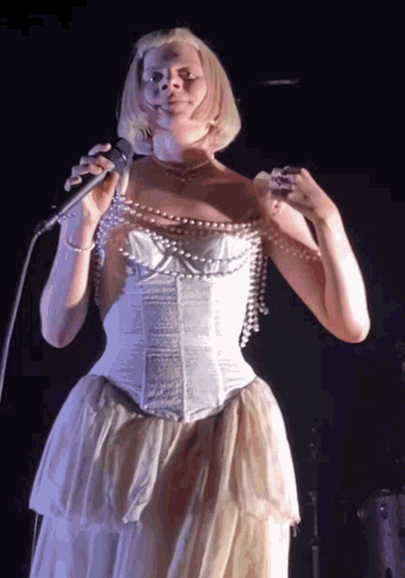 a woman in a white dress is singing into a microphone with pearls around her neck