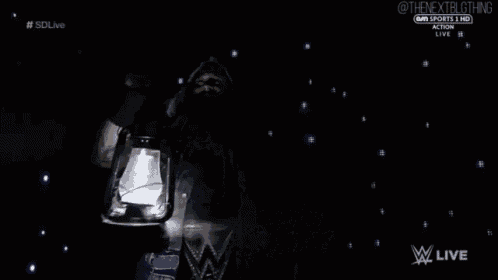 a wrestler is holding a lantern in the dark while wearing a hood .