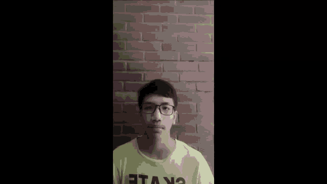 a young man wearing glasses and a yellow shirt that says " ekvile " is standing in front of a brick wall