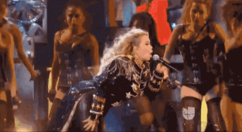 a woman is singing into a microphone while a group of dancers are dancing around her .