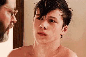 a shirtless young man is looking at himself in the mirror while a man looks on .