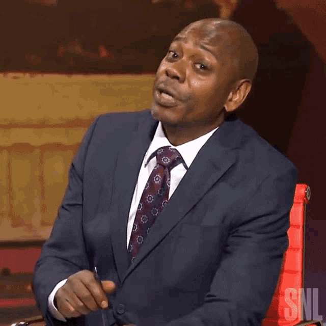 a man in a suit and tie is sitting in a red chair with the snl logo on the bottom