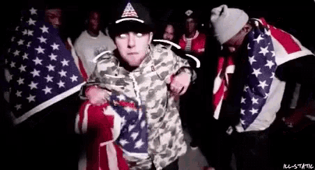 a man in a camo jacket is holding an american flag in front of a group of people .