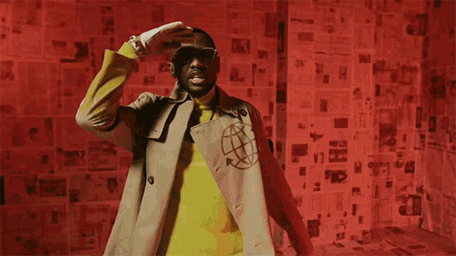 a man wearing sunglasses and a trench coat stands in front of a wall covered in newspapers