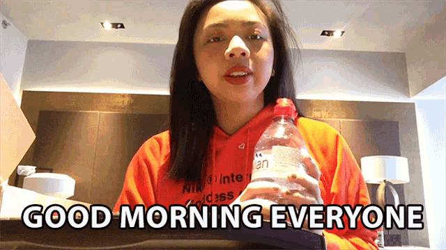 a woman in an orange hoodie is holding a bottle of water and saying good morning everyone