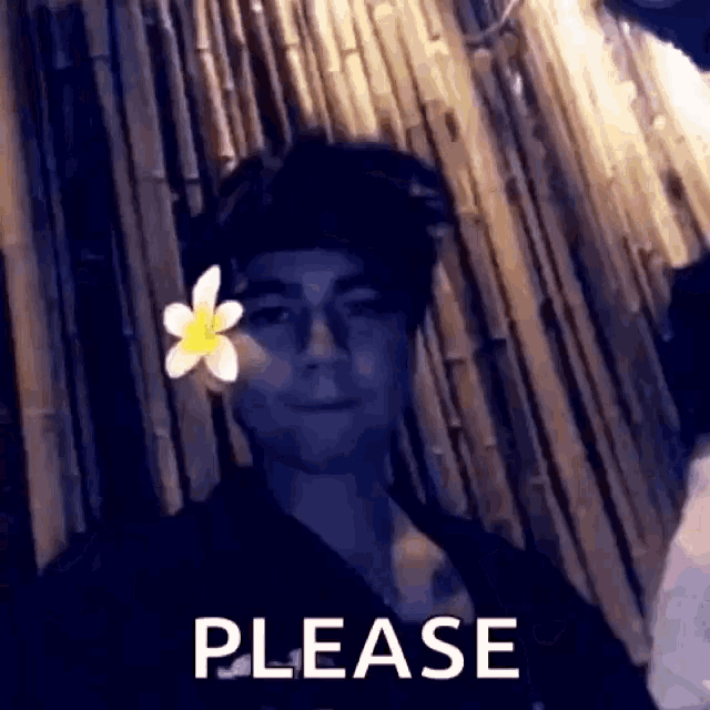 a man with a flower in his hair is standing in front of a bamboo wall and says please .