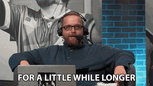 a man wearing headphones says " for a little while longer " while sitting in front of a laptop