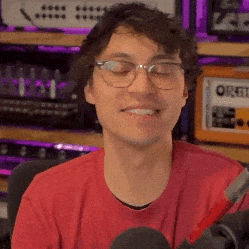 a young man wearing glasses and a red shirt is smiling while sitting in front of a microphone .