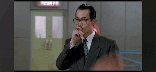 a man in a suit and tie is smoking a cigarette in front of a red exit sign .