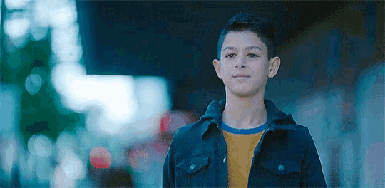 a young boy wearing a black jacket and a yellow t-shirt is standing on a city street .