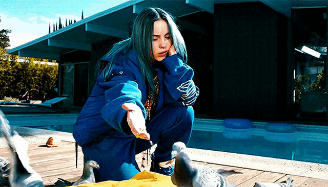 a woman in a blue jacket is feeding pigeons near a swimming pool