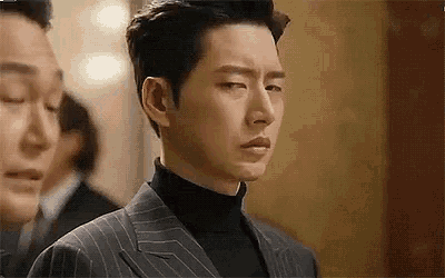 a man in a suit and black turtleneck is standing next to another man in a suit .