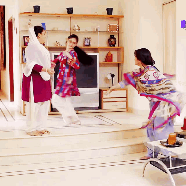 three women are dancing in a living room with a tv in the background