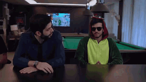 two men are sitting at a table in front of a pool table and one of them is wearing a green dinosaur costume