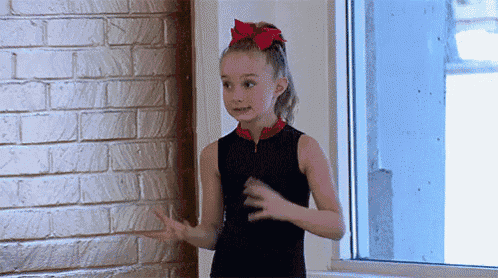 a young girl with a red bow in her hair is standing in front of a brick wall .