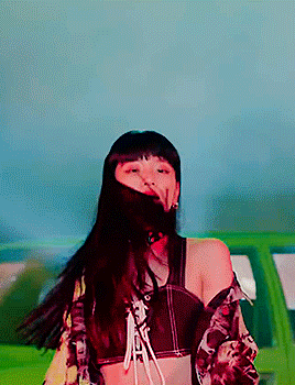 a woman in a crop top is standing in front of a green car with her hair blowing in the wind .
