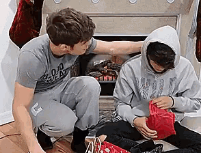 a man in a gray shirt is kneeling next to another man in a hoodie who is holding a red gift box .