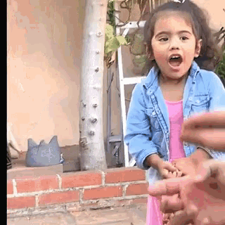 a little girl in a pink dress and denim jacket is holding someone 's hand and making a funny face .
