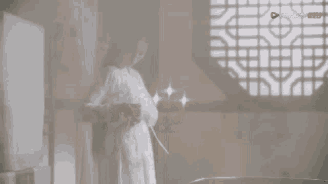 a woman in a white robe is standing in front of a window holding a candle .