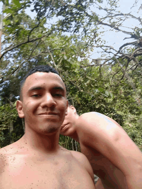 a shirtless man takes a selfie with another shirtless man behind him
