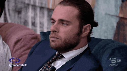 a man in a suit and tie is sitting on a couch in front of a screen that says grandefratello