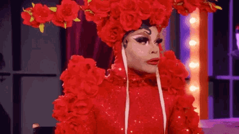 a drag queen is wearing a red costume with red flowers on her head .