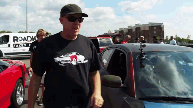 a man wearing a black shirt that says roadk takes stands in front of a car