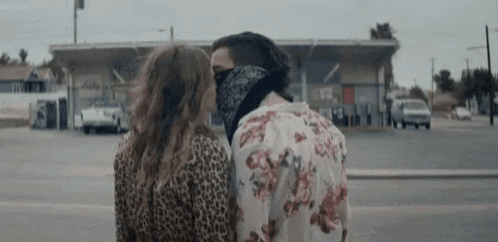 a man and a woman are kissing in front of a gas station . the man is wearing a bandana on his face .