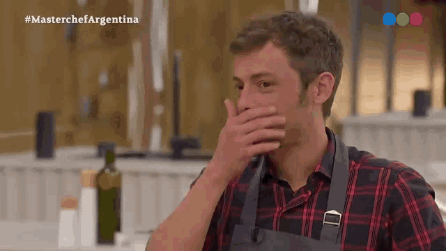 a man in a plaid shirt and apron is covering his mouth with his hand in a kitchen .