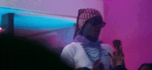 a man wearing a bandana and sunglasses is standing in front of a pink wall and holding a cell phone .