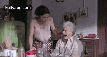 an elderly woman is sitting at a table with a woman standing next to her .