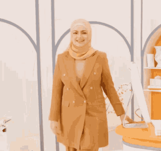 a woman wearing a hijab and a tan suit is standing in front of a desk