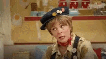 a woman wearing a hat and overalls is making a face .