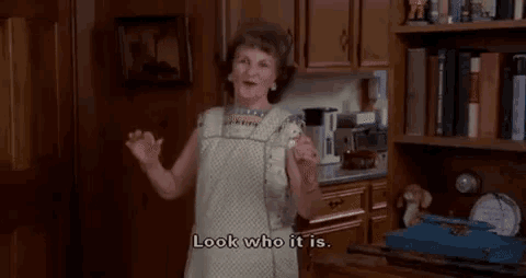 an elderly woman is standing in a kitchen holding an ice cream cone and saying `` look who it is . ''