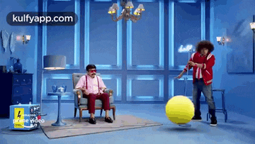 a man in a pink shirt is sitting in a chair while a man in a red jacket is spinning a yellow ball