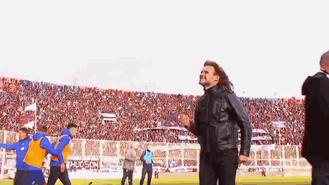 a man in a leather jacket stands on a soccer field in front of a crowd