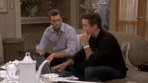 two men are sitting on a couch looking at laptops