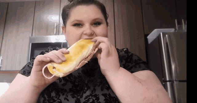 a woman in a black shirt is eating a sandwich in a kitchen