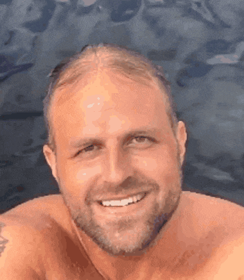 a shirtless man with a beard is smiling in front of a body of water .