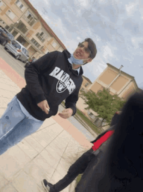 a man wearing a hoodie that says raiders is standing on a sidewalk