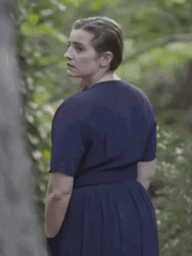 a woman in a blue dress is standing in the woods and looking at something .