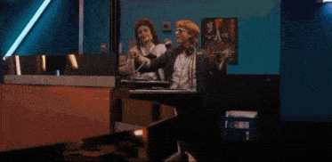 a man with curly hair is making a funny face while sitting next to another man in a dark room .