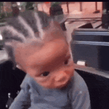 a baby with braids on his head is sitting in a stroller and looking at the camera .