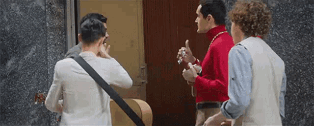 three men are standing in front of a door talking to each other . one man is holding a guitar .