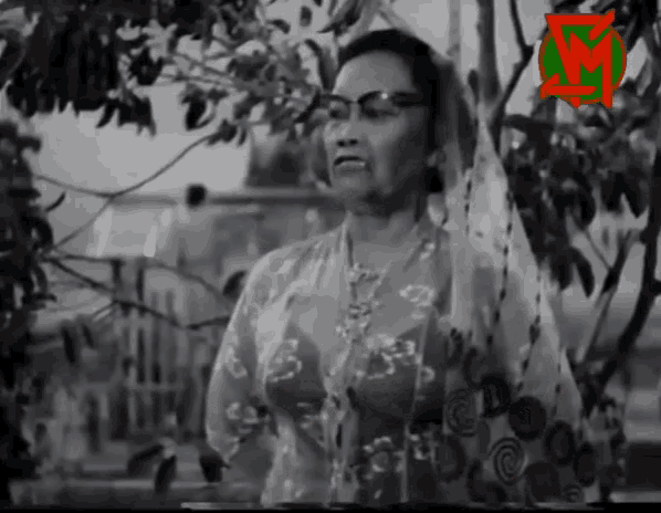 a woman wearing glasses stands in front of a tree with the letter m in the corner