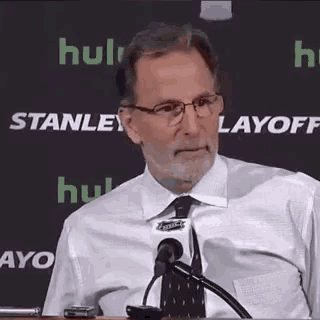 a man wearing glasses and a tie is sitting at a podium talking into a microphone .