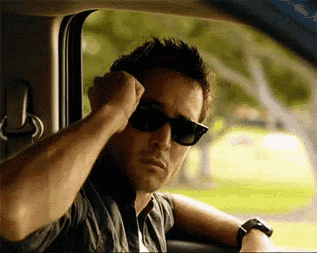 a man wearing sunglasses sits in a car with his hand on his head