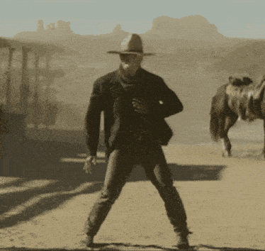 a man in a cowboy hat is standing in the desert with a horse behind him